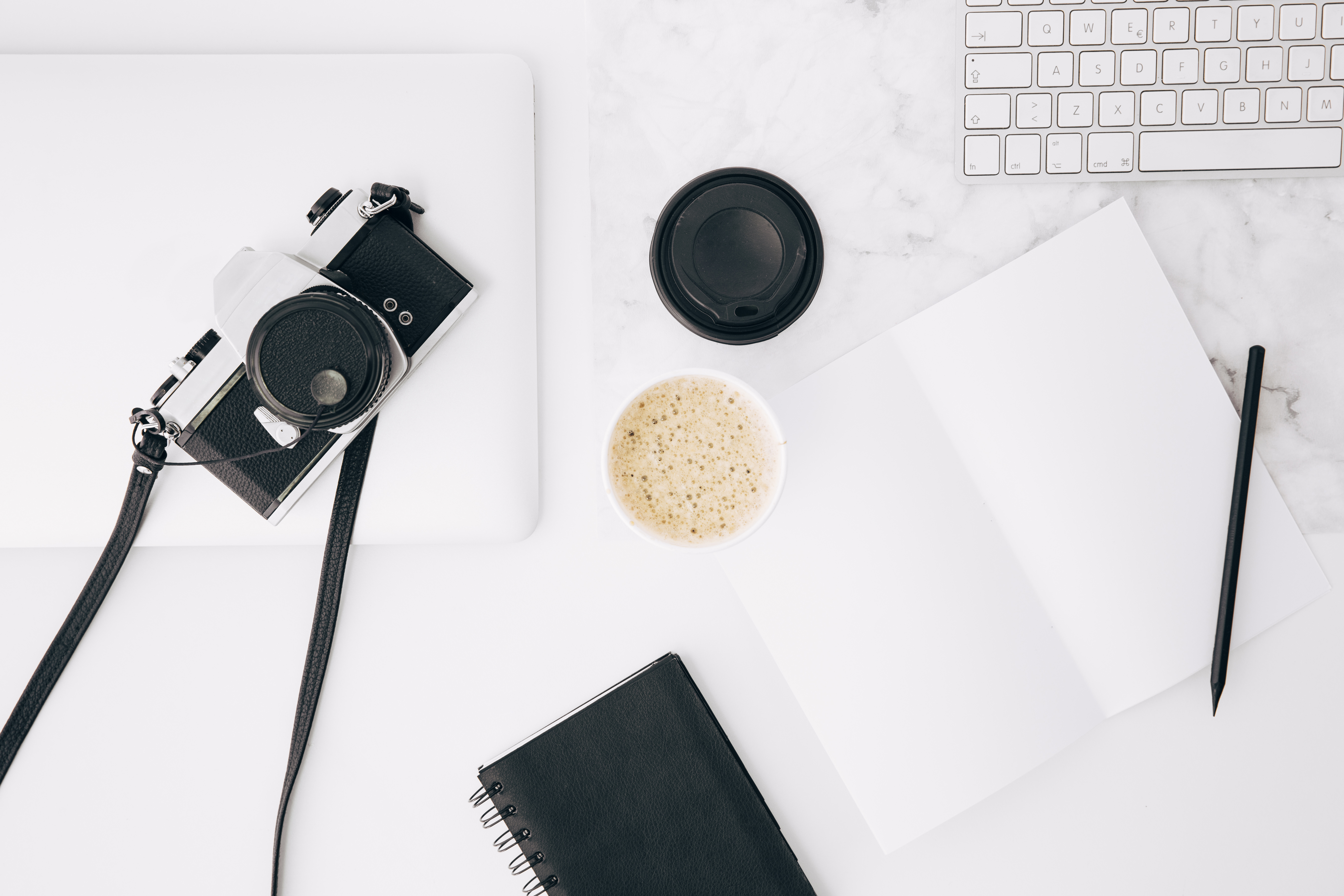 White working desk with a retro camera, notebook, pen, keyboard, and coffee.